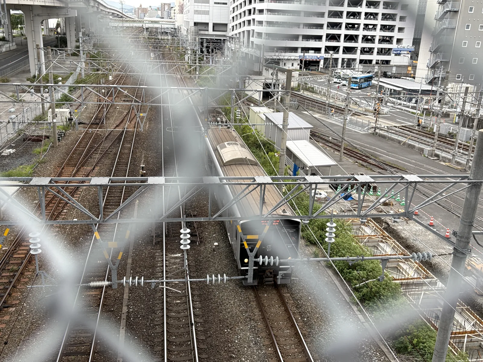 ランプの上からフェンス越しに見下ろしたJRの線路。電車が走り、右奥にコムシティの立体駐車場が見える