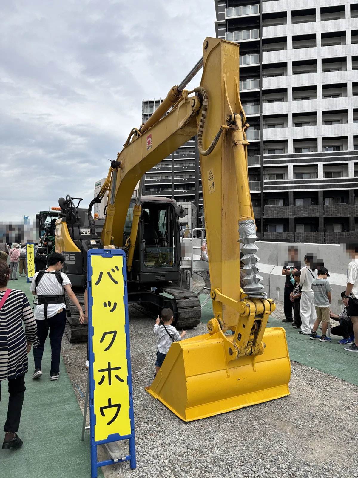 「バックホウ」の看板と、黄色いバックホウのアーム。足もとで子どもが見上げている
